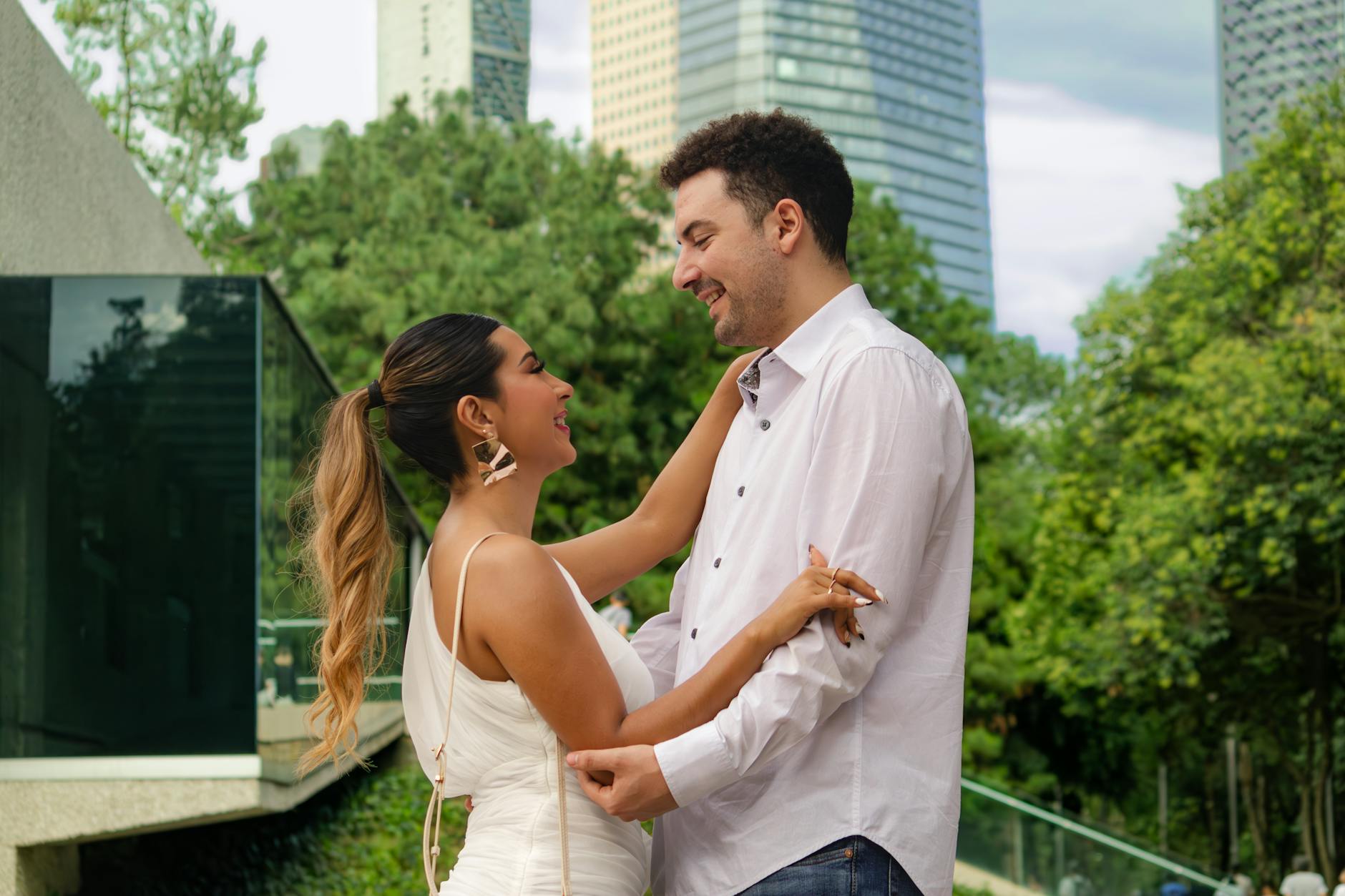 A smiling couple shares a tender moment in a lush park in Ciudad de México, with modern city buildings in the background.