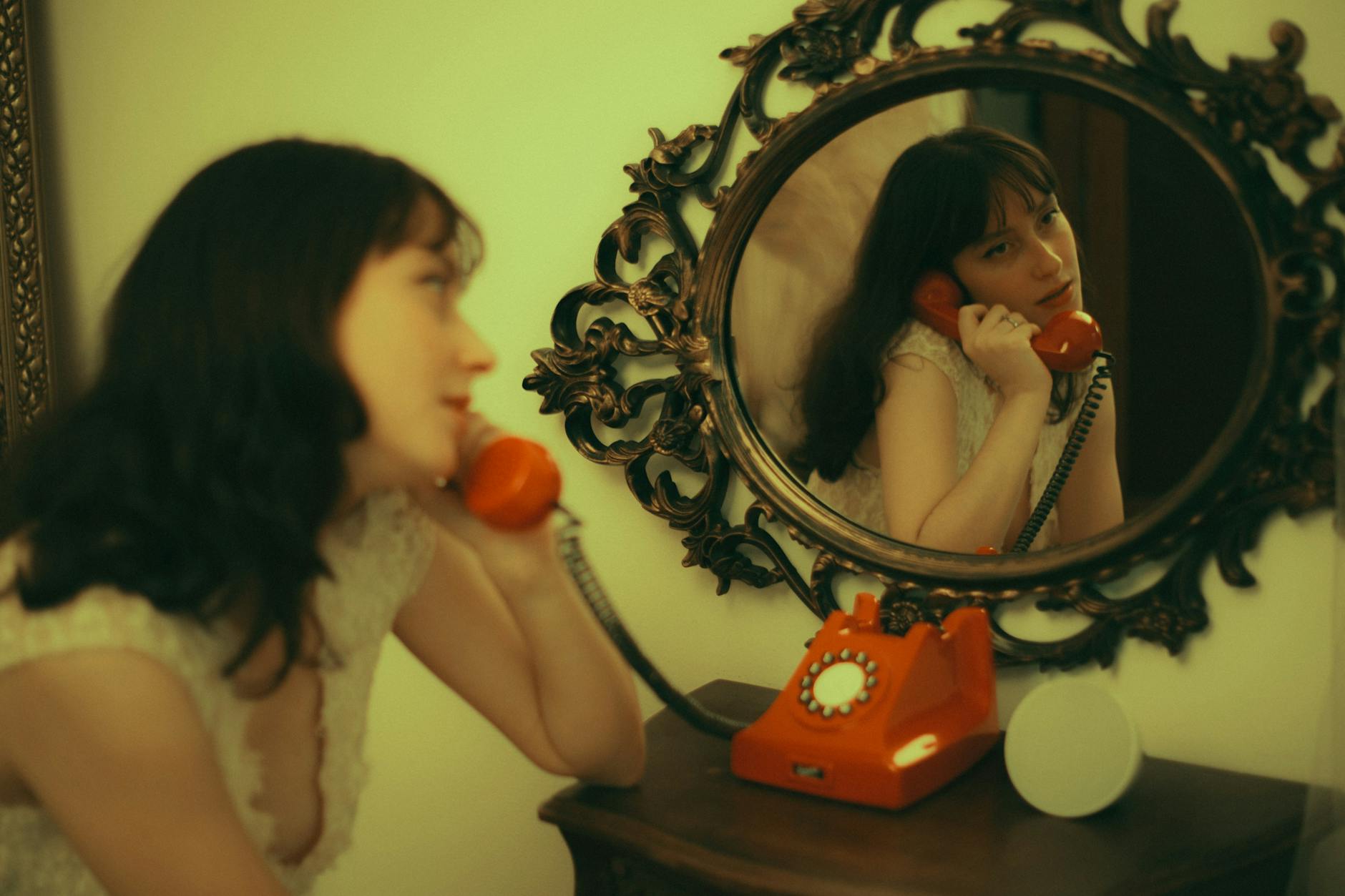 Artistic vintage-style photo of a woman using an orange rotary phone in the mirror.