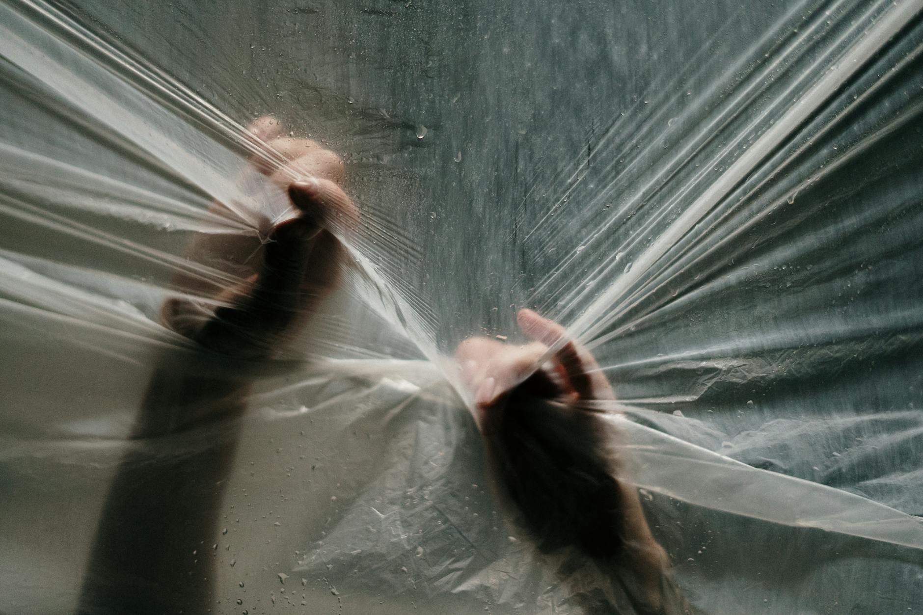Hands gripping and stretching a transparent plastic film with water droplets and light effects.