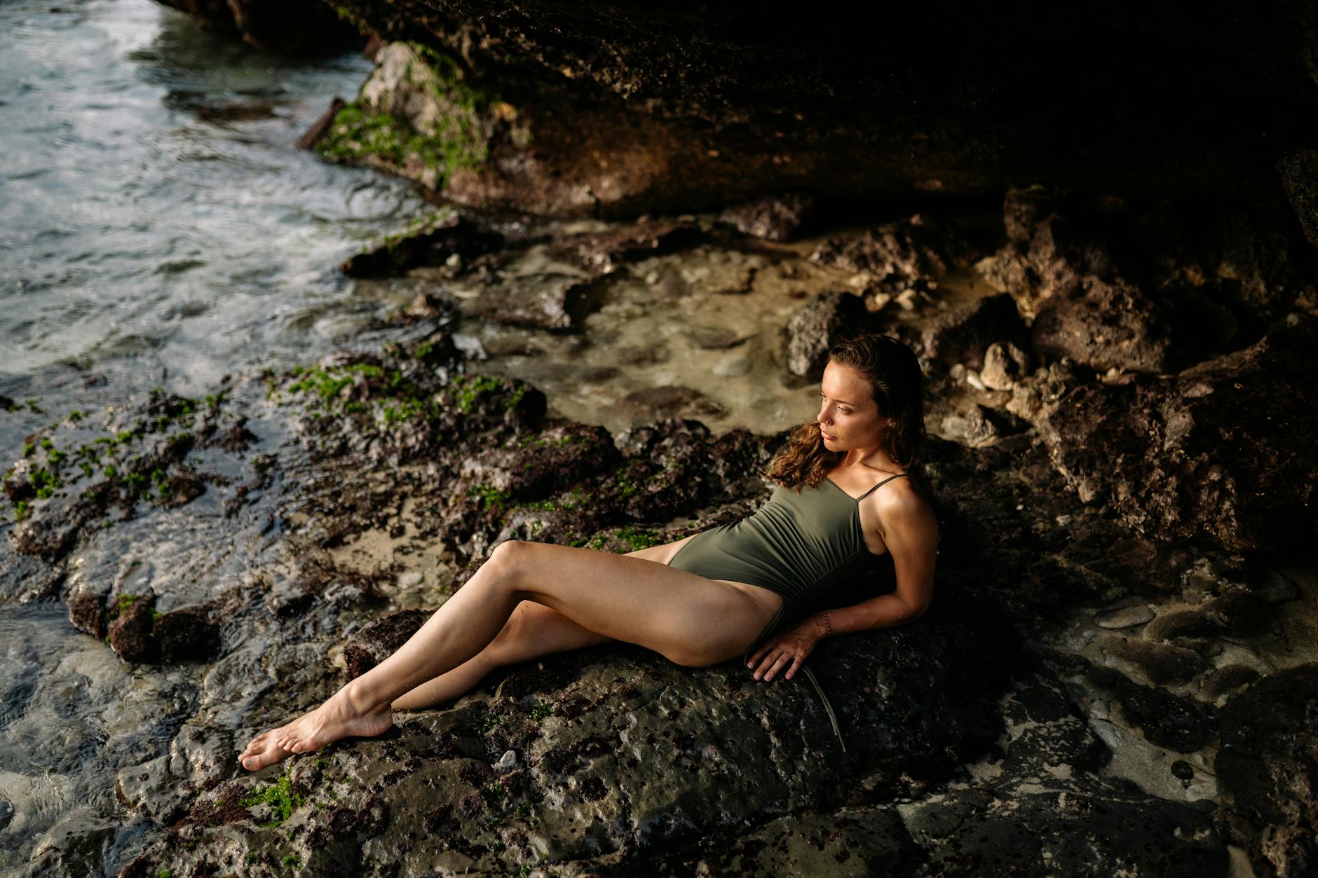 A serene woman in a swimsuit relaxes on rocky shore by the ocean at sunset.
