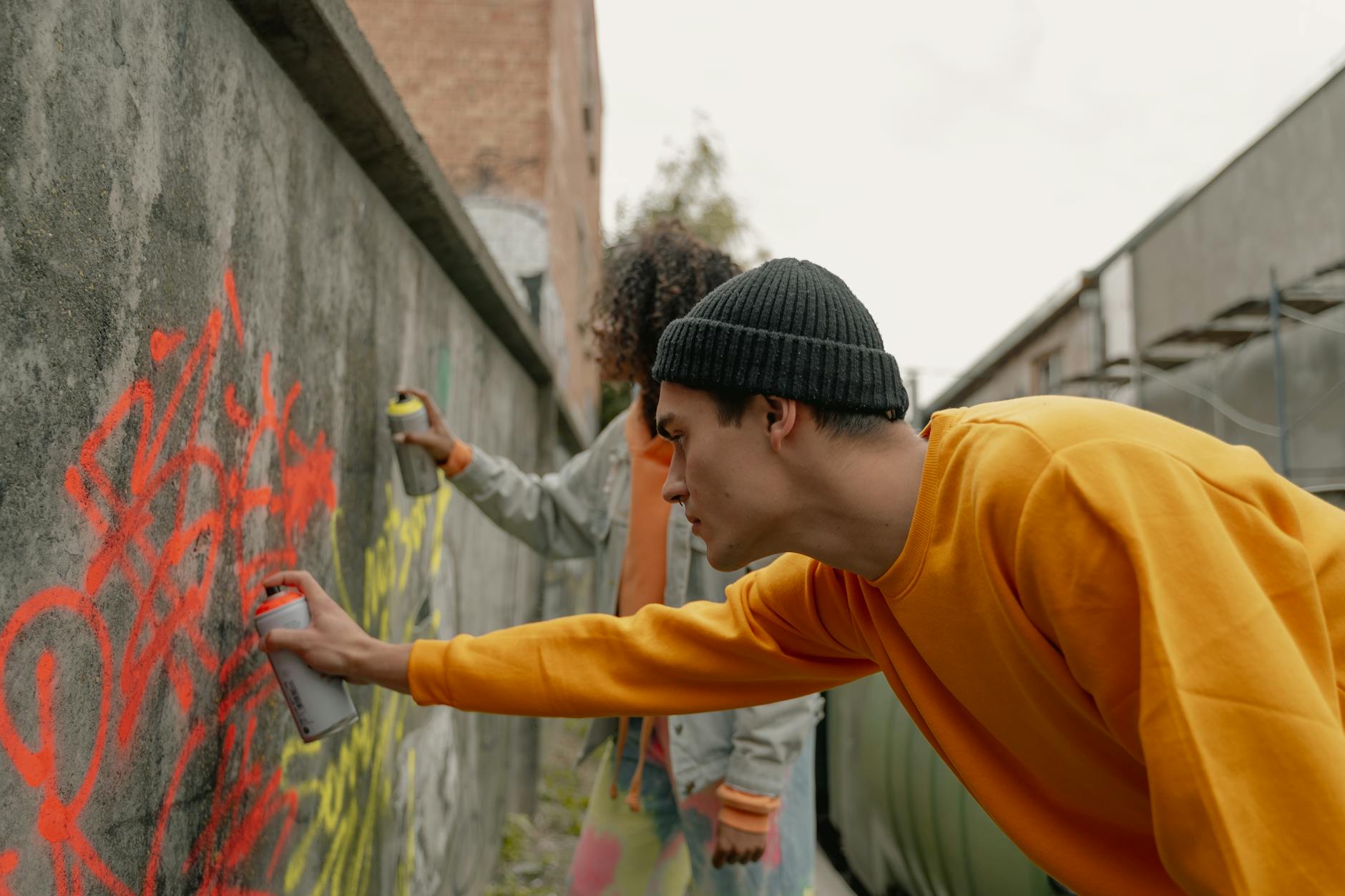 Two adults spray painting colorful graffiti on a concrete wall in an urban setting.