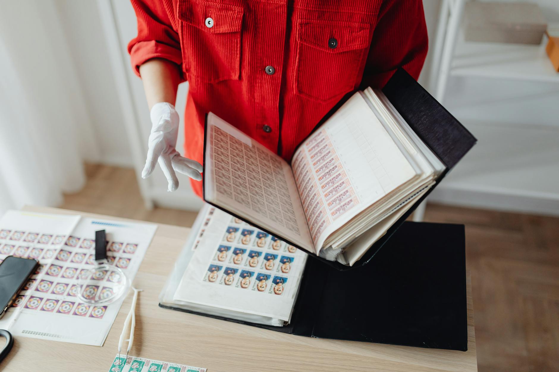 Stamp collector in red shirt browsing stamp album indoors with magnifying glass.