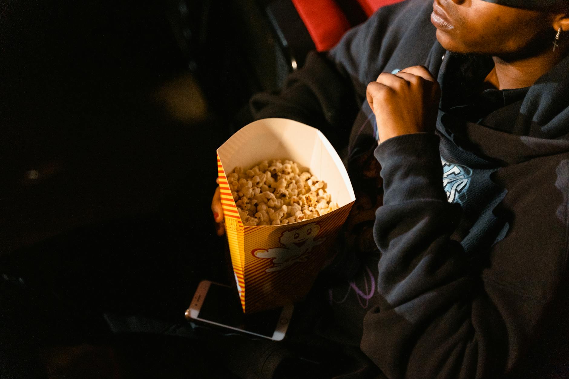 A person holds popcorn while watching a film in a dark movie theater.