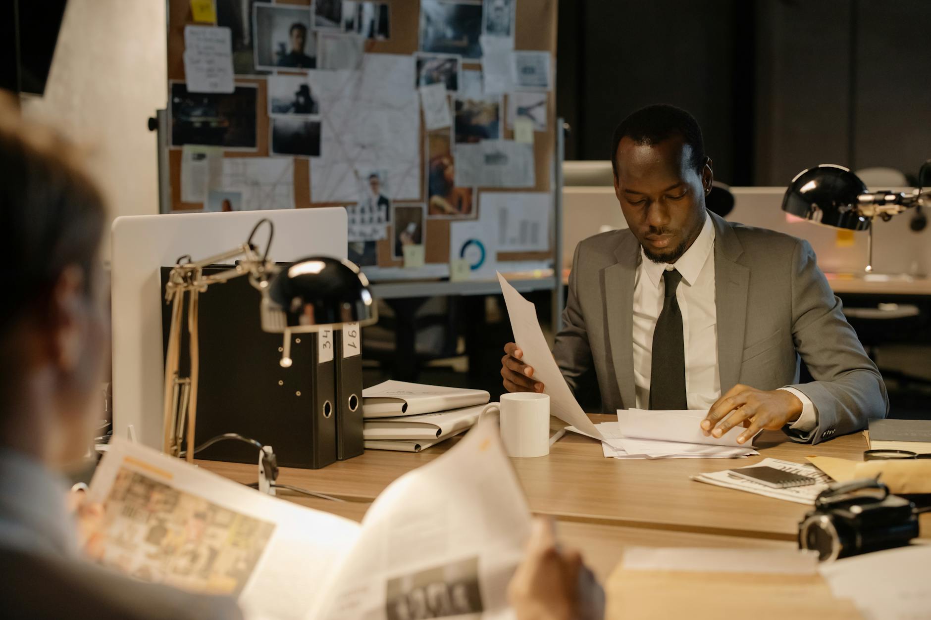 Detective in a suit examines papers at desk in a dimly-lit office, suggesting investigation work.