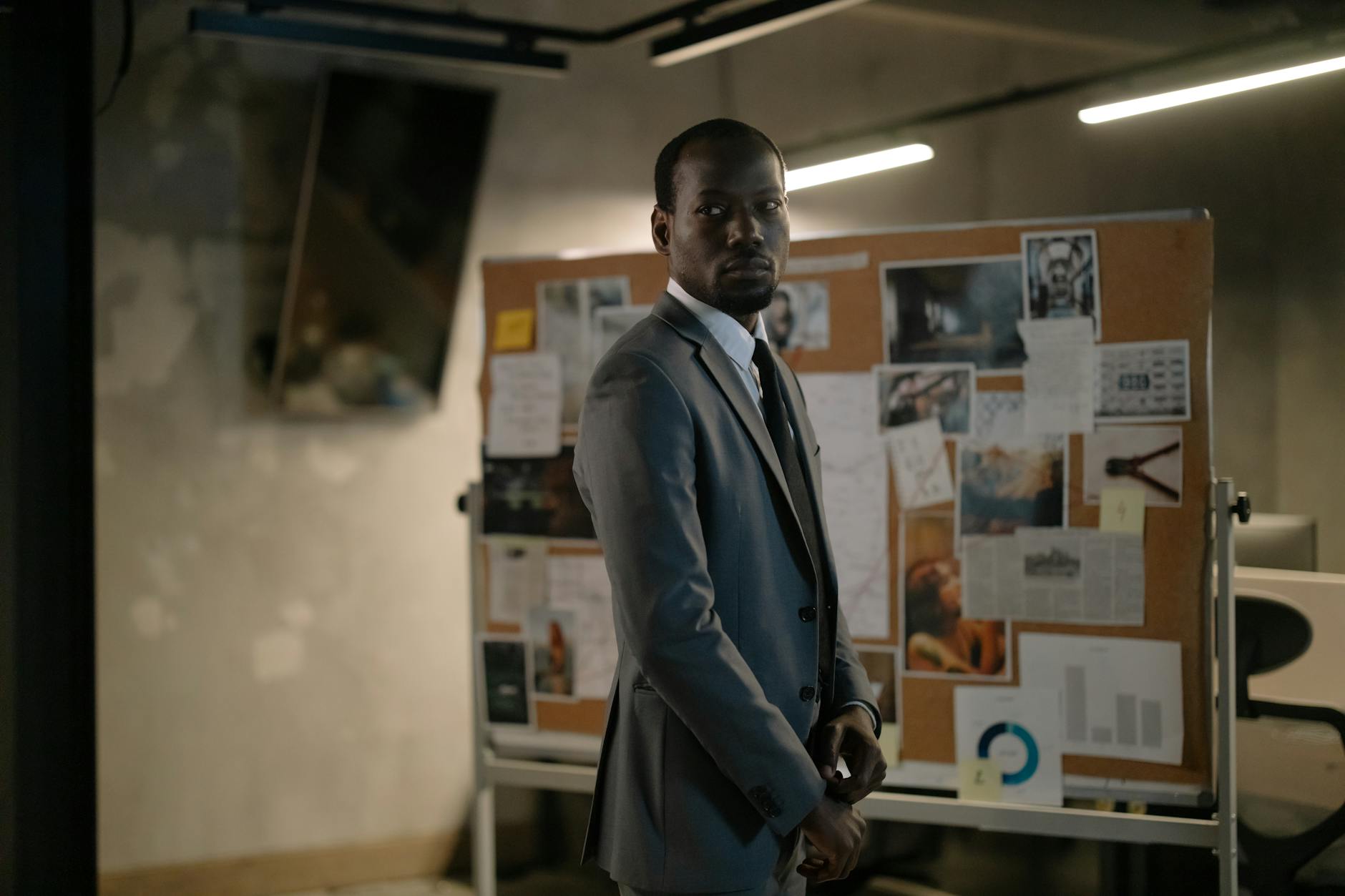 A serious man in a suit stands in an office, examining a bulletin board with notes and photos.