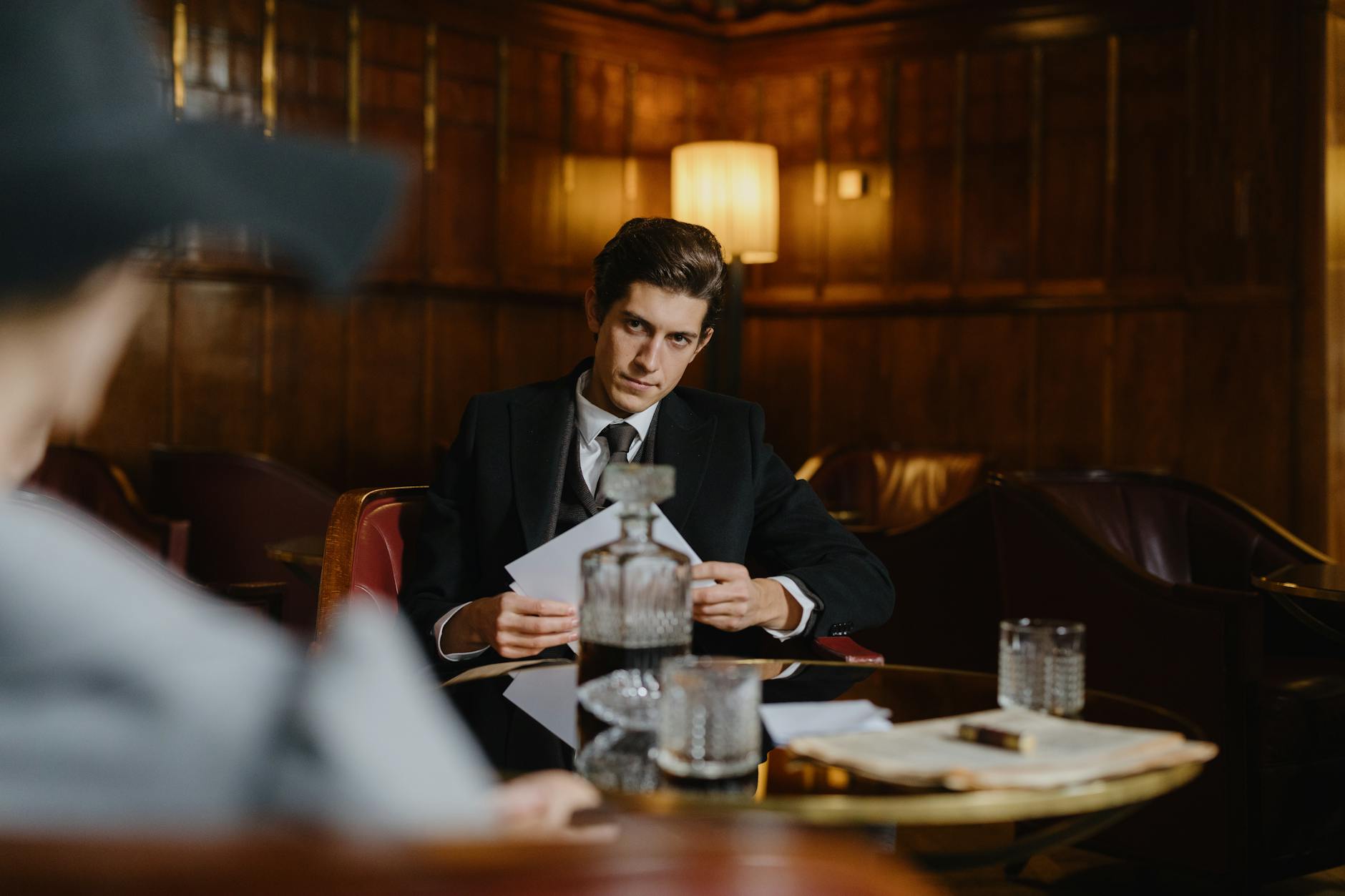 A serious man in a suit sitting at a table with papers, evoking a corporate or mobster vibe in an elegant, dimly lit room.