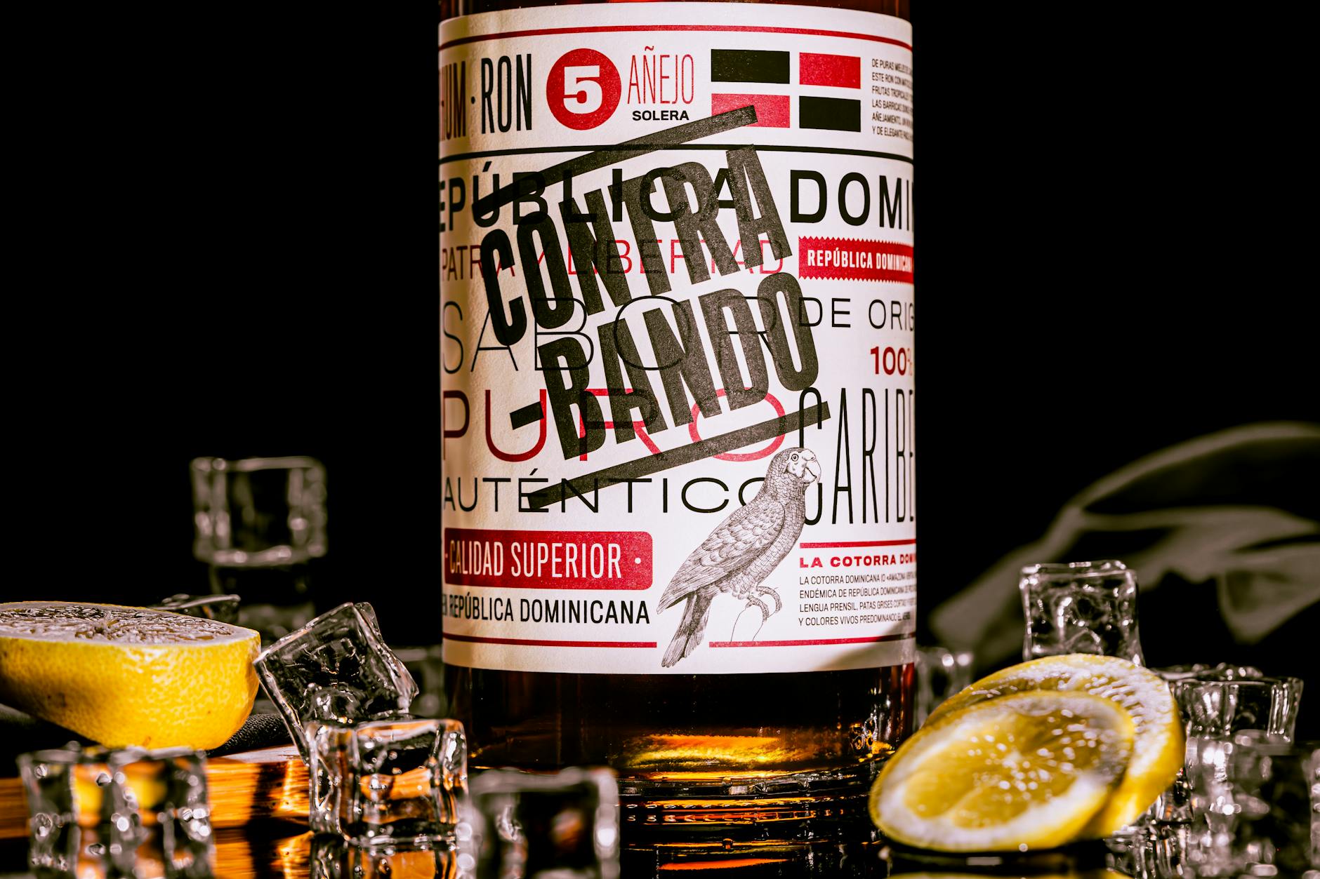 Close-up of Contrabando rum bottle with lemon slices and ice cubes on a reflective surface.