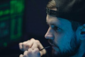 A young man in a cap biting on wire, deeply focused, with glowing tech-screen ambiance.