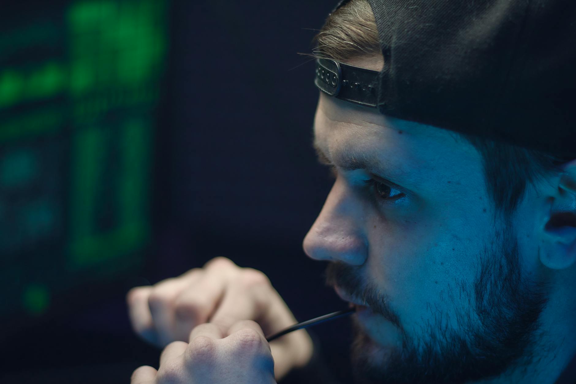 A young man in a cap biting on wire, deeply focused, with glowing tech-screen ambiance.